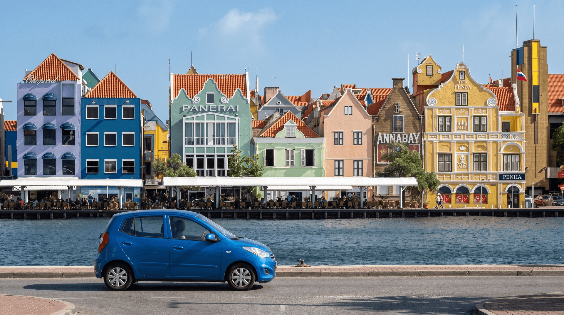 Small blue rental hatchback driving along the colorful Handelskade waterfront in Willemstad, Curaçao