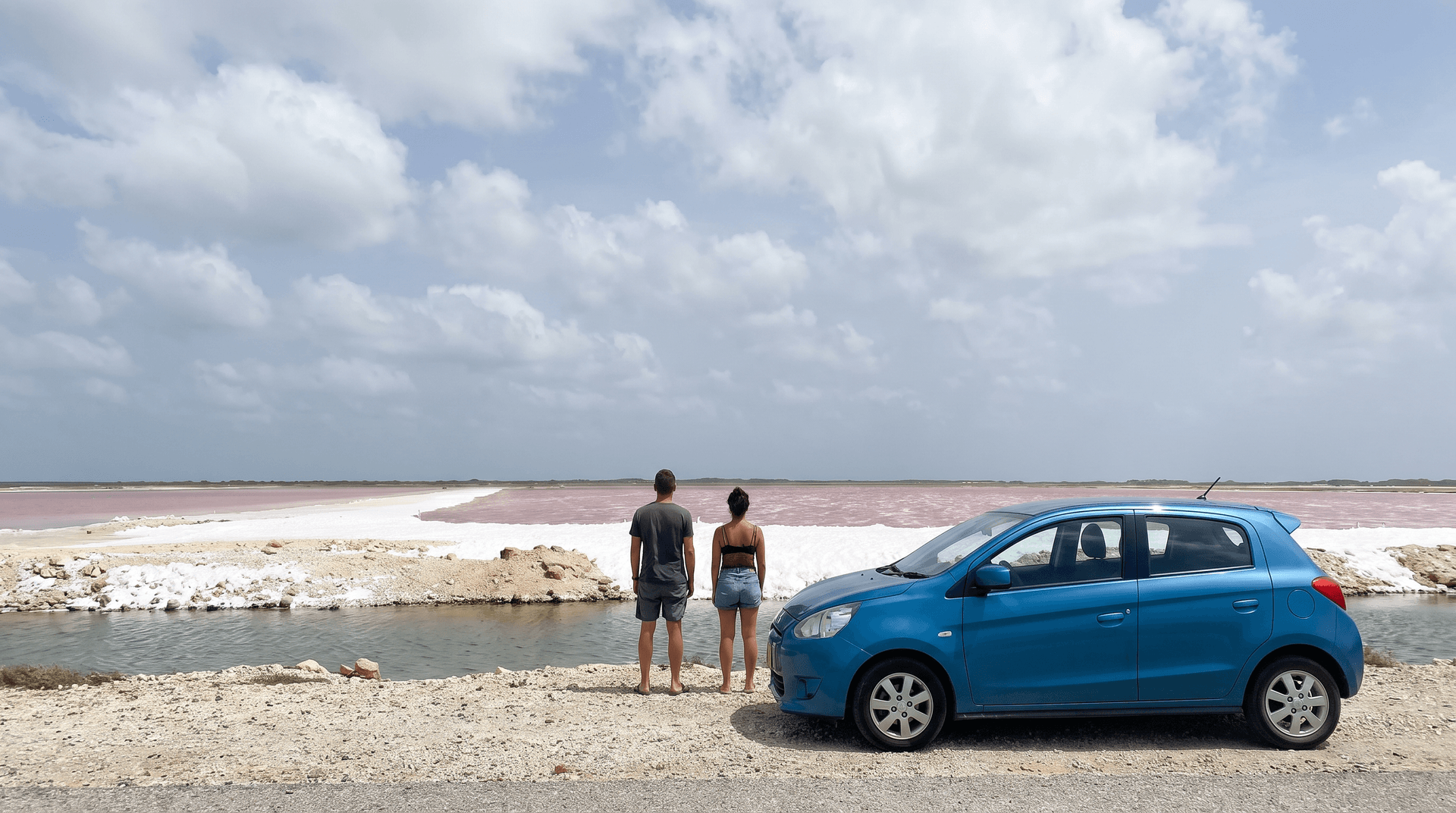 Two travelers beside a blue hatchback looking at the pink salt pans in southern Bonaire